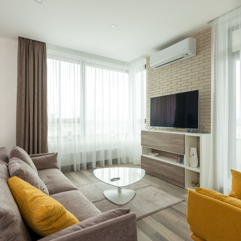 intérieur salon moderne avec climatiseur réversible mural blanc installé au-dessus d'un meuble TV, canapé gris avec coussins jaune moutarde.