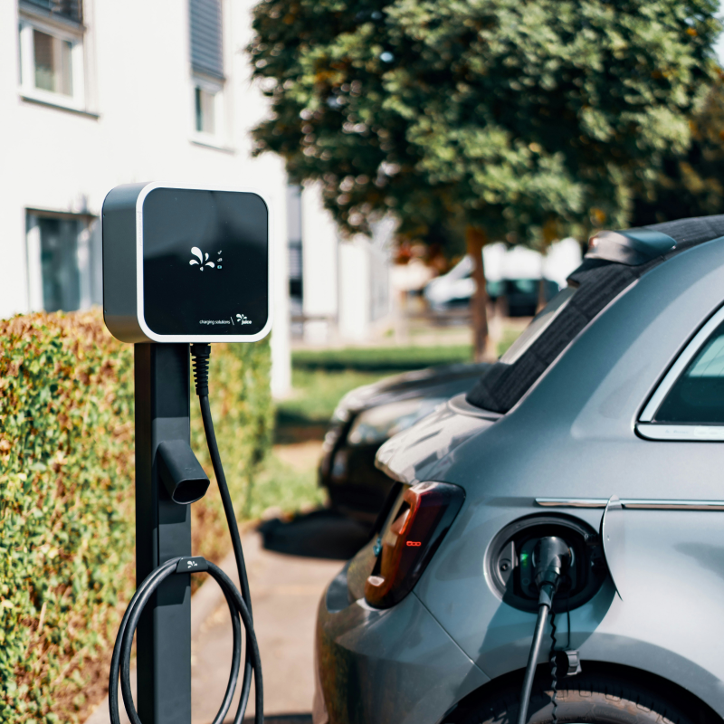 Borne de recharge murale pour véhicule électrique installée sur pied, rechargeant une voiture grise dans une allée résidentielle.